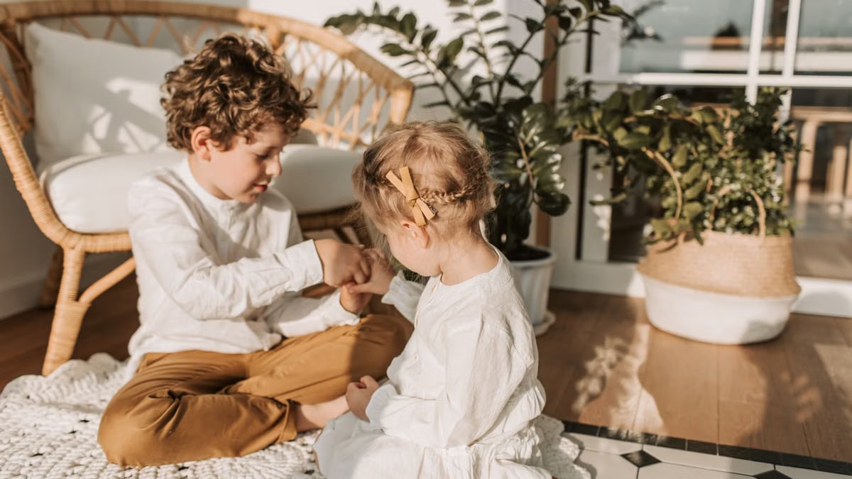 Two siblings sit together on a rug in a bright, plant-filled room, enjoying a playful moment.